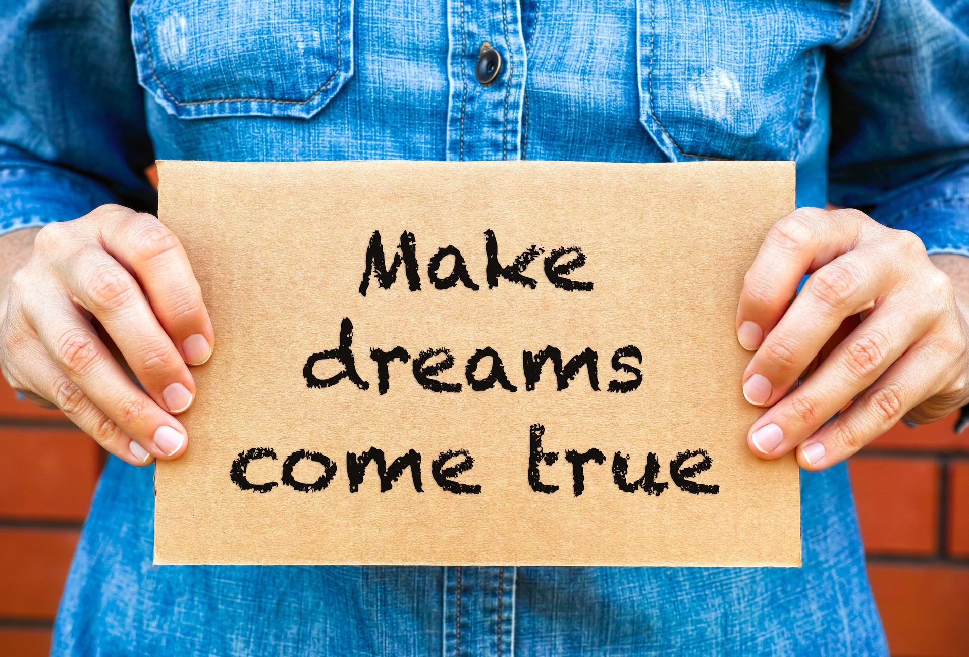 A woman holding a piece of a cardboard with words Make Dreams Come True against a brick wall background.